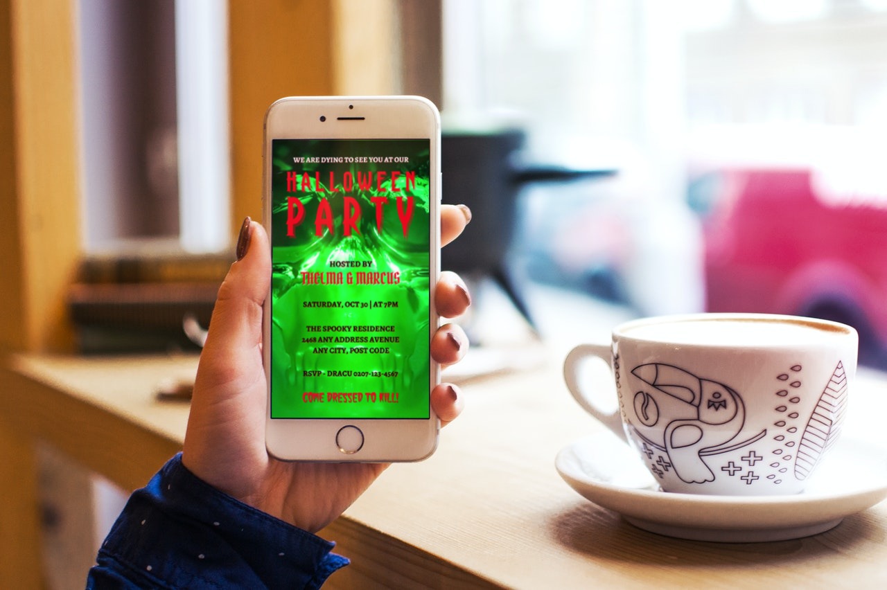 Hand holding a phone with a green screen, showcasing the Neon Green Skull Halloween Party Video Invite from URCordiallyInvited.