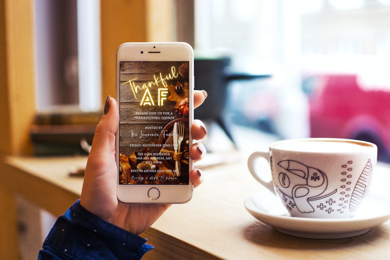 Hand holding a phone displaying Thankful AF Neon Wooden Table | Thanksgiving Dinner Evite invitation template, with a cup of coffee in the background.