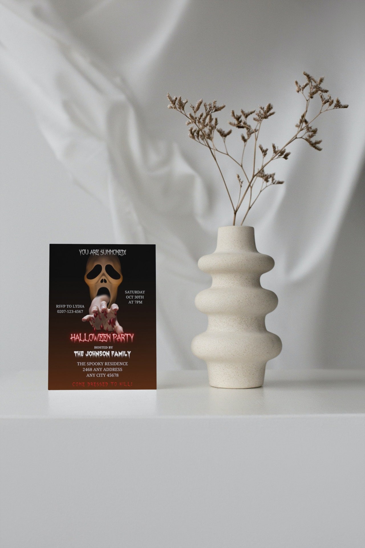 White vase with dried flowers beside a Halloween invitation card featuring a hand with blood, titled Scary Mouth Hand Skull | Halloween Evite.
