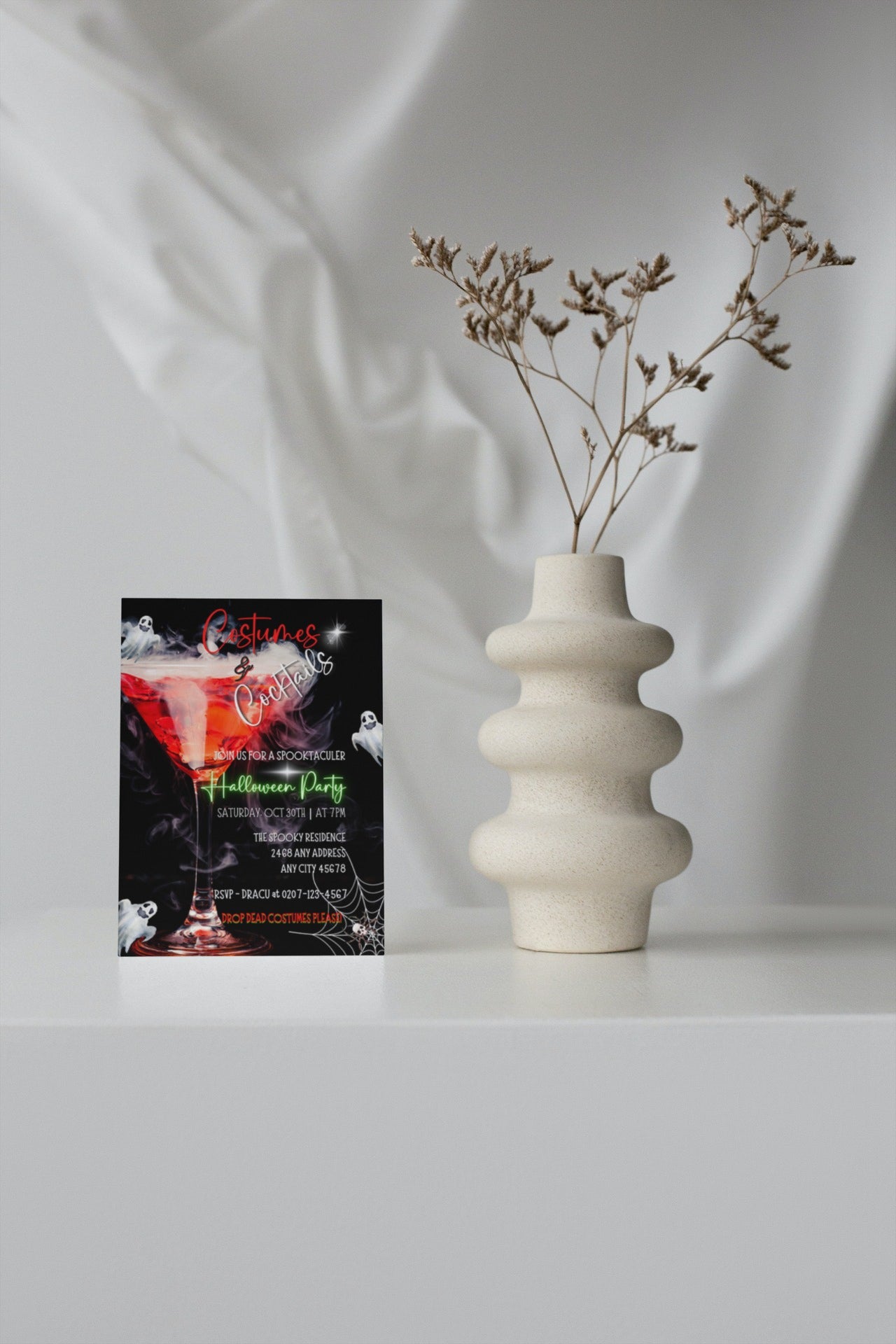 White vase with dried flowers next to Halloween party invitation card featuring a smoking wine glass, ghost costumes, and cocktails graphic.