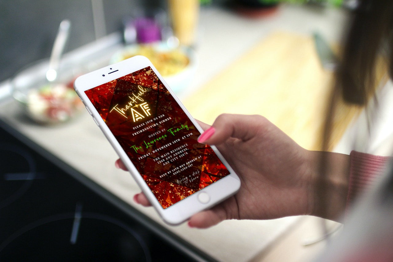 Hand holding a phone displaying the Rustic Red Gold Leafy | Thankful AF Thanksgiving Evite template on its screen.