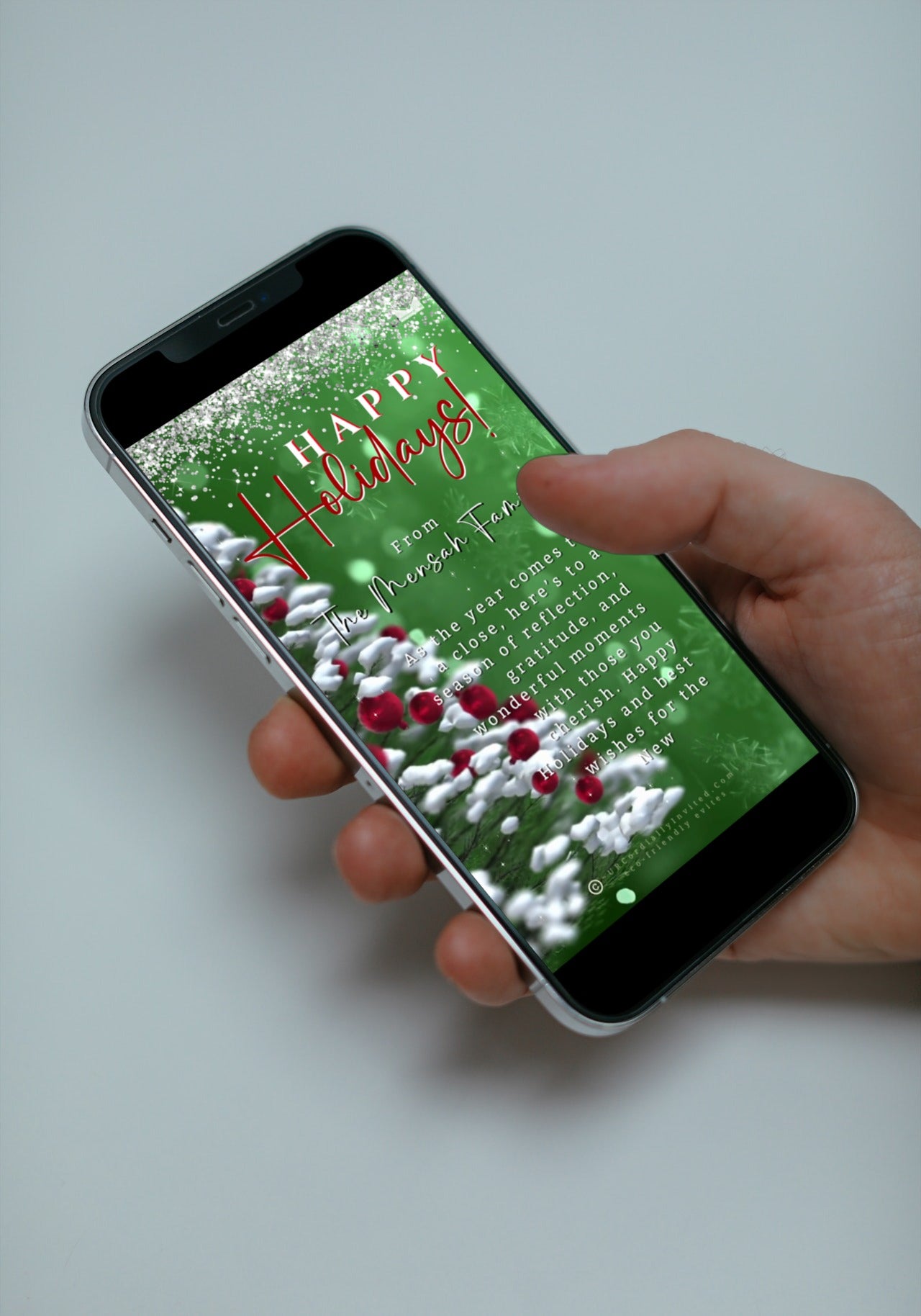 Hand holding a smartphone displaying a Snow Crystals Green White Christmas Video Greeting Ecard, customizable via Canva for digital invitations.