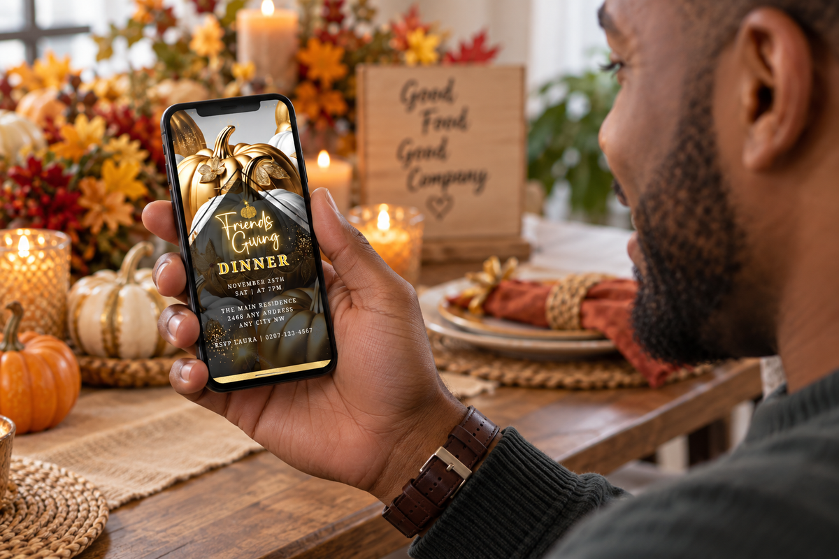 Hand holding phone displaying gold pumpkin Friendsgiving invitation; festive autumn table decor in background.