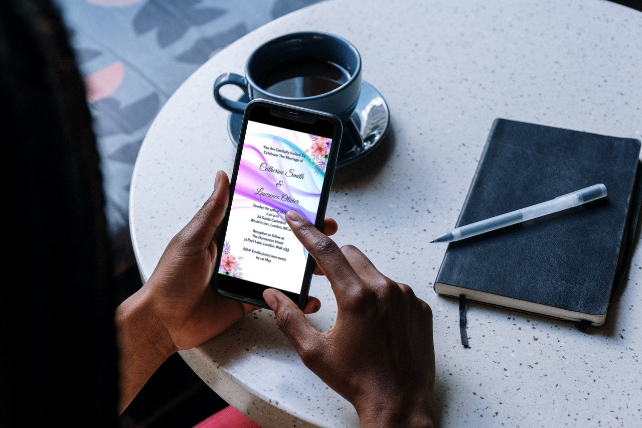 Person holding a smartphone displaying a customizable Lilac White Floral Wedding Video Invitation template from URCordiallyInvited. Pen and book visible in the background.