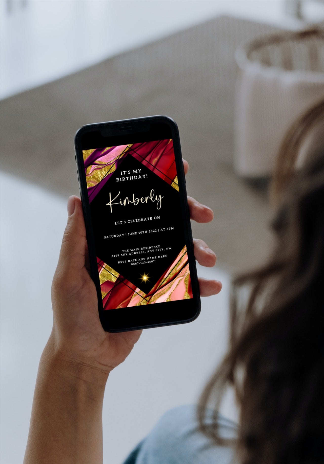 A hand holding a smartphone displaying a Maroon Ankara Themed Editable Birthday Evite template for customization via Canva.