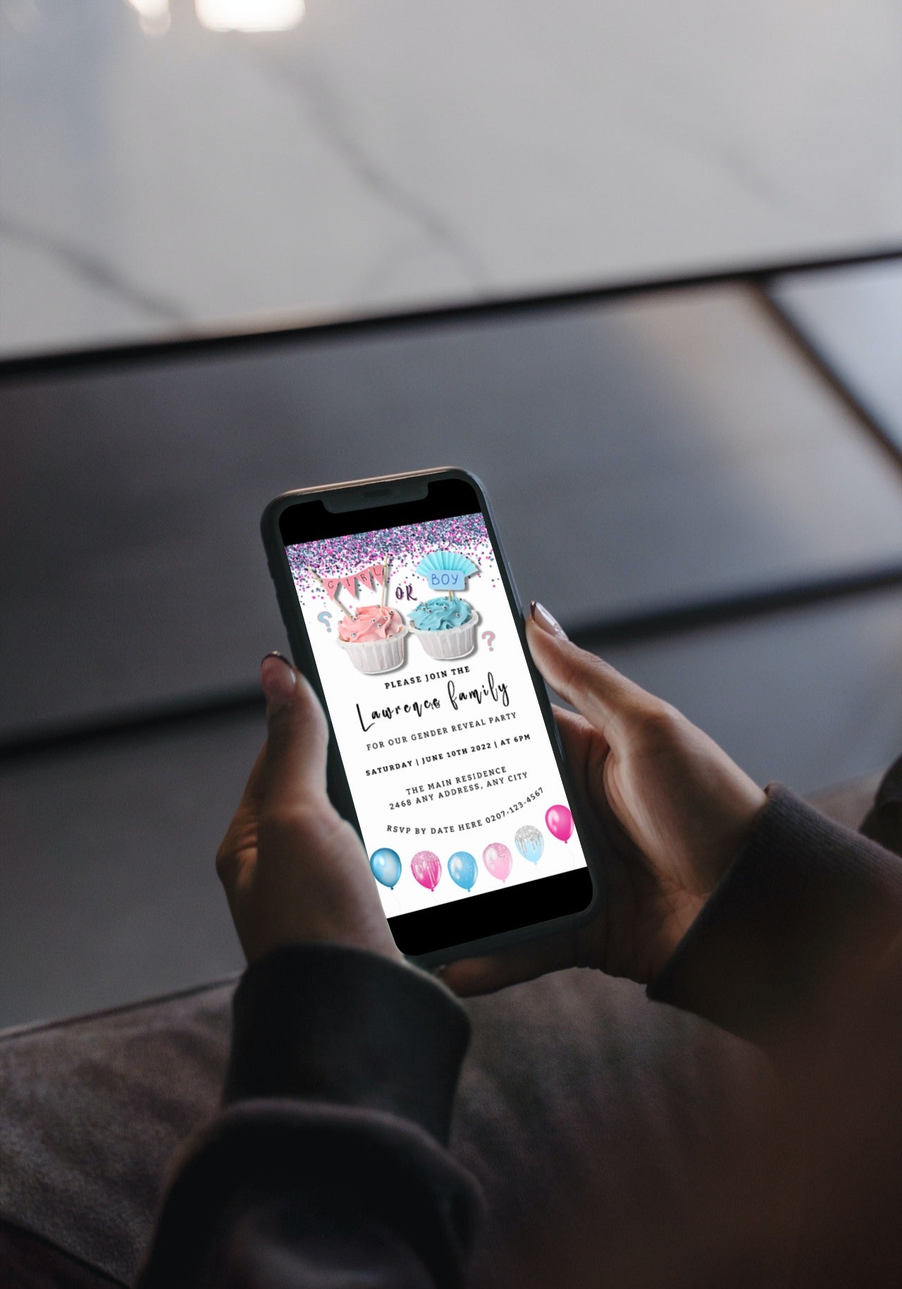 Person holding a smartphone displaying a customizable digital invitation for a gender reveal with cupcakes and balloons.
