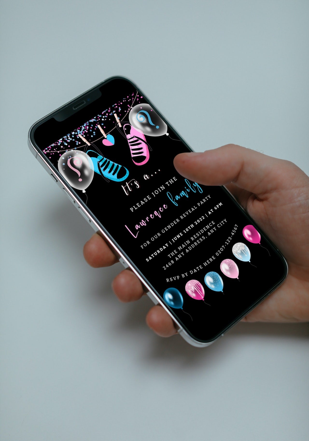 Hand holding a smartphone displaying customizable digital invitation with black, blue, and pink baby shoes for a gender reveal event.