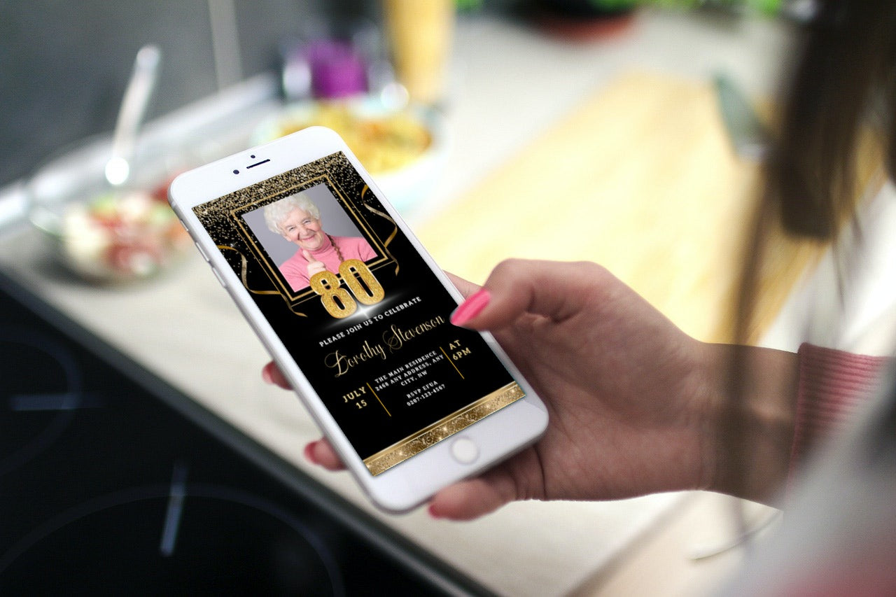 Hand holding a smartphone displaying a customisable 80th Birthday Evite with black gold confetti and a photo of an elderly woman.