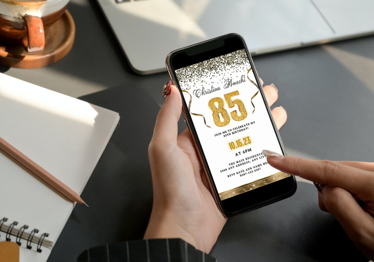 Person holding a smartphone displaying the customisable White Gold Confetti 85th Birthday Evite template from URCordiallyInvited, ready for personalisation in Canva.