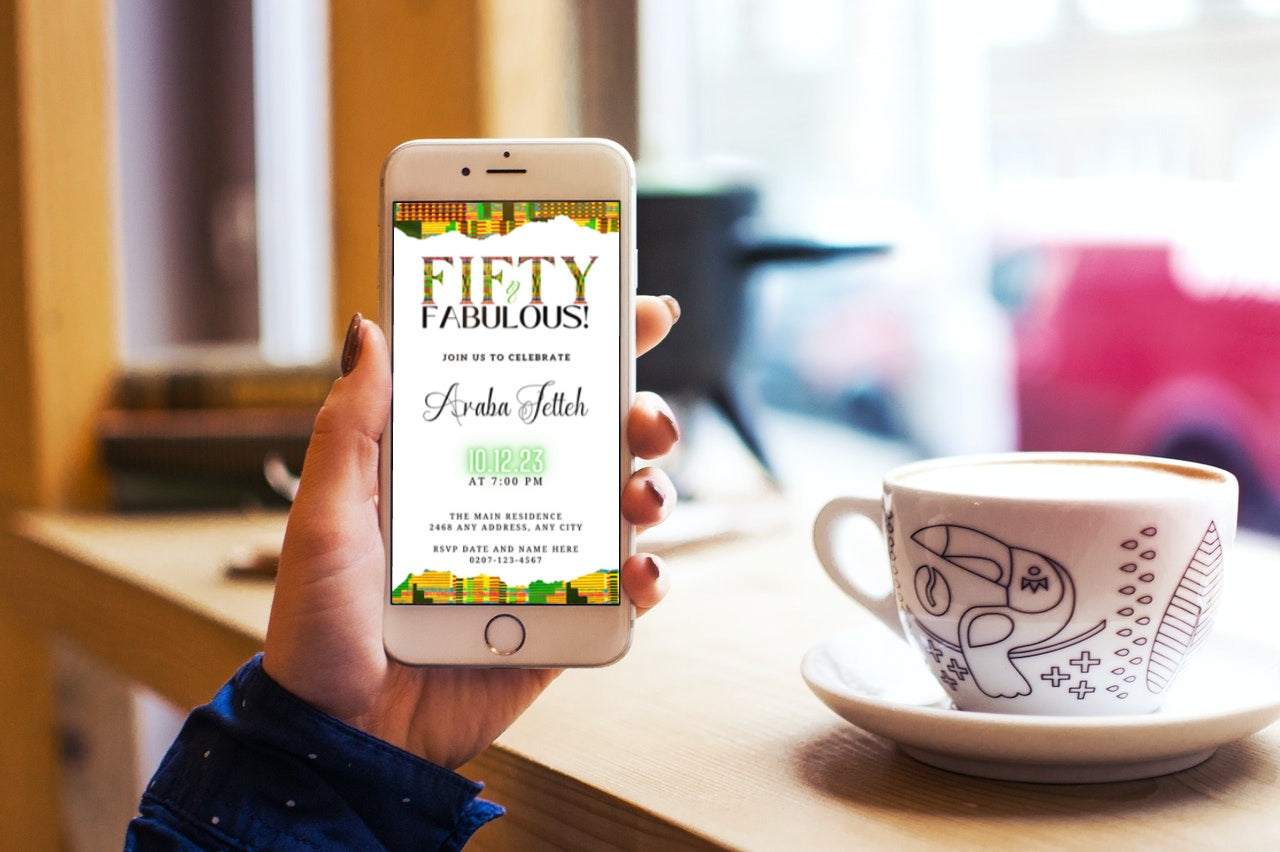 Hand holding a phone displaying the customizable Green Yellow Kente White | 50 & Fabulous Party Evite template, with a cup of coffee nearby.