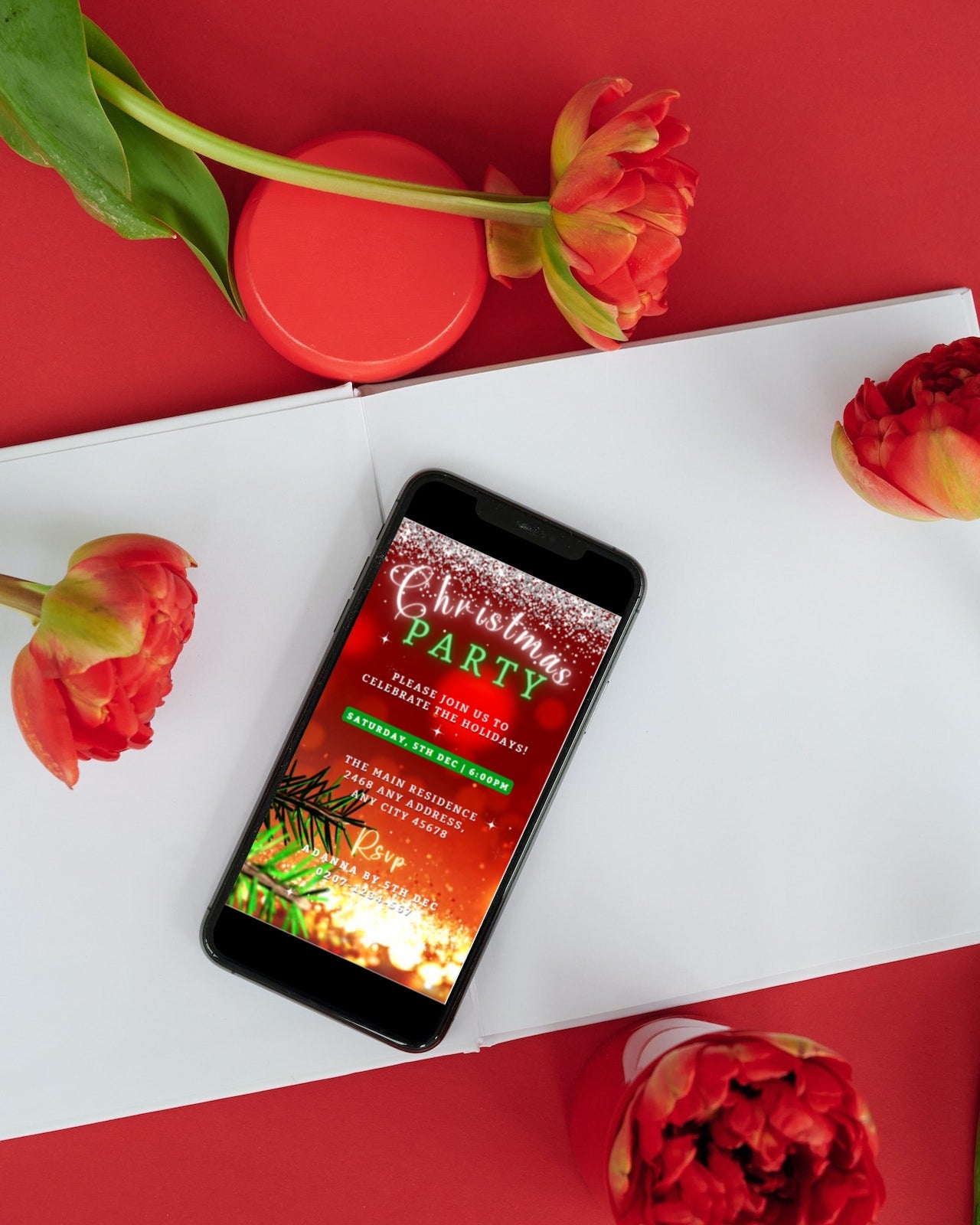 Phone on a table with flowers, showcasing the Red Gold Glitter Christmas Video Invitation customizable via Canva for smartphone use.