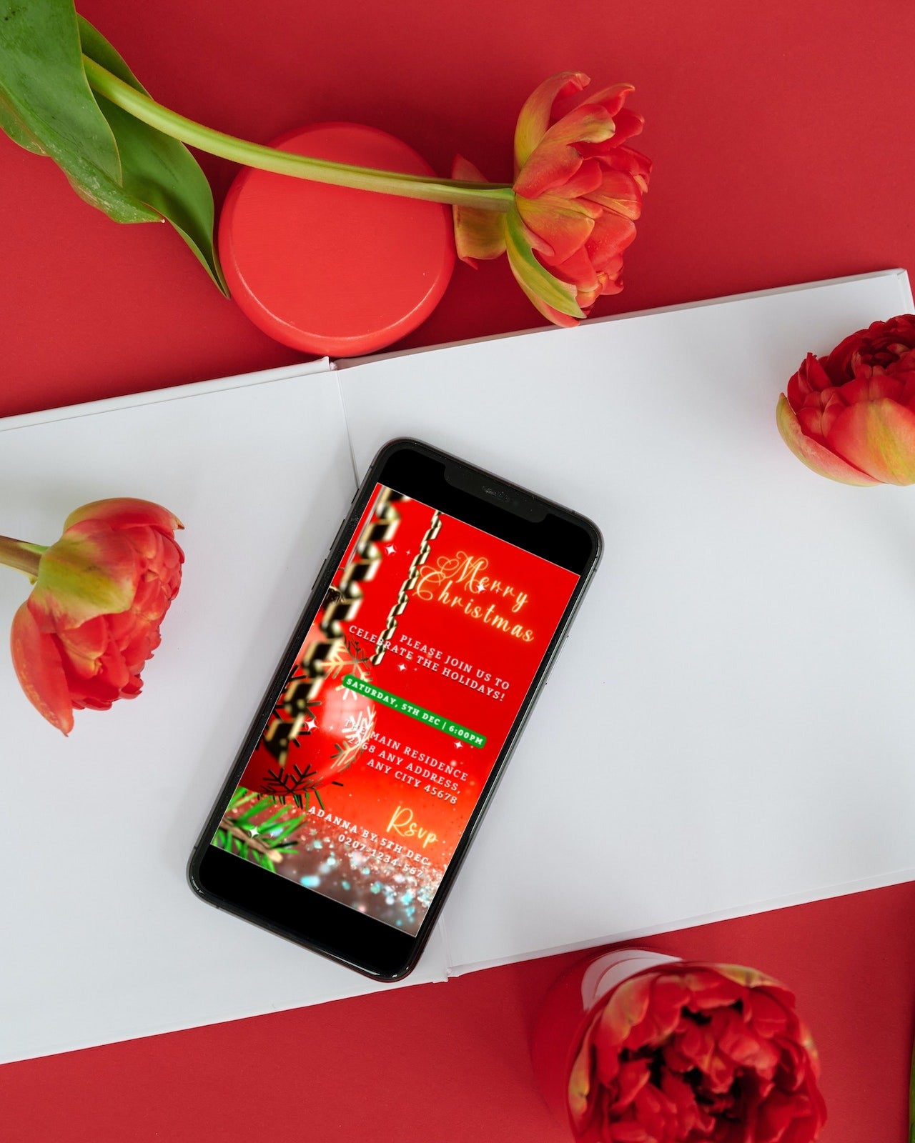 Smartphone displaying editable Red Gold Ornament Glitter Christmas video invitation template, accompanied by flowers and a book, emphasizing the customizable ecard for events.