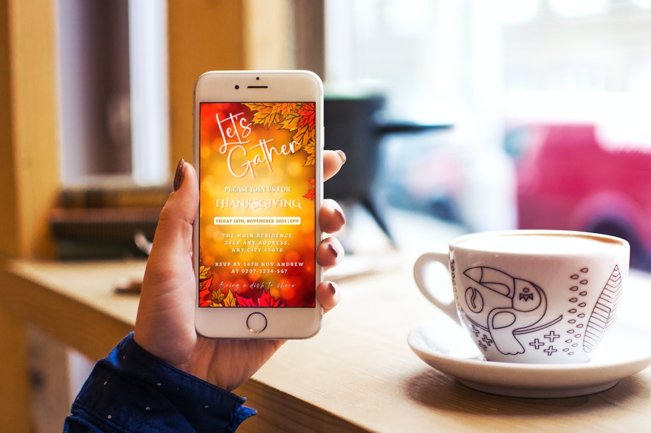 Hand holding a cell phone displaying the Let's Gather Orange Leaves Bokeh | Thanksgiving Dinner Evite invitation template for customization via Canva.