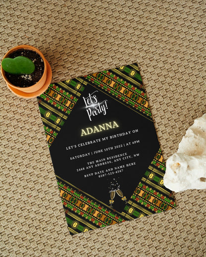 Black and gold Green Black African Ankara invitation on a carpet, accompanied by a green potted plant and a white rock.