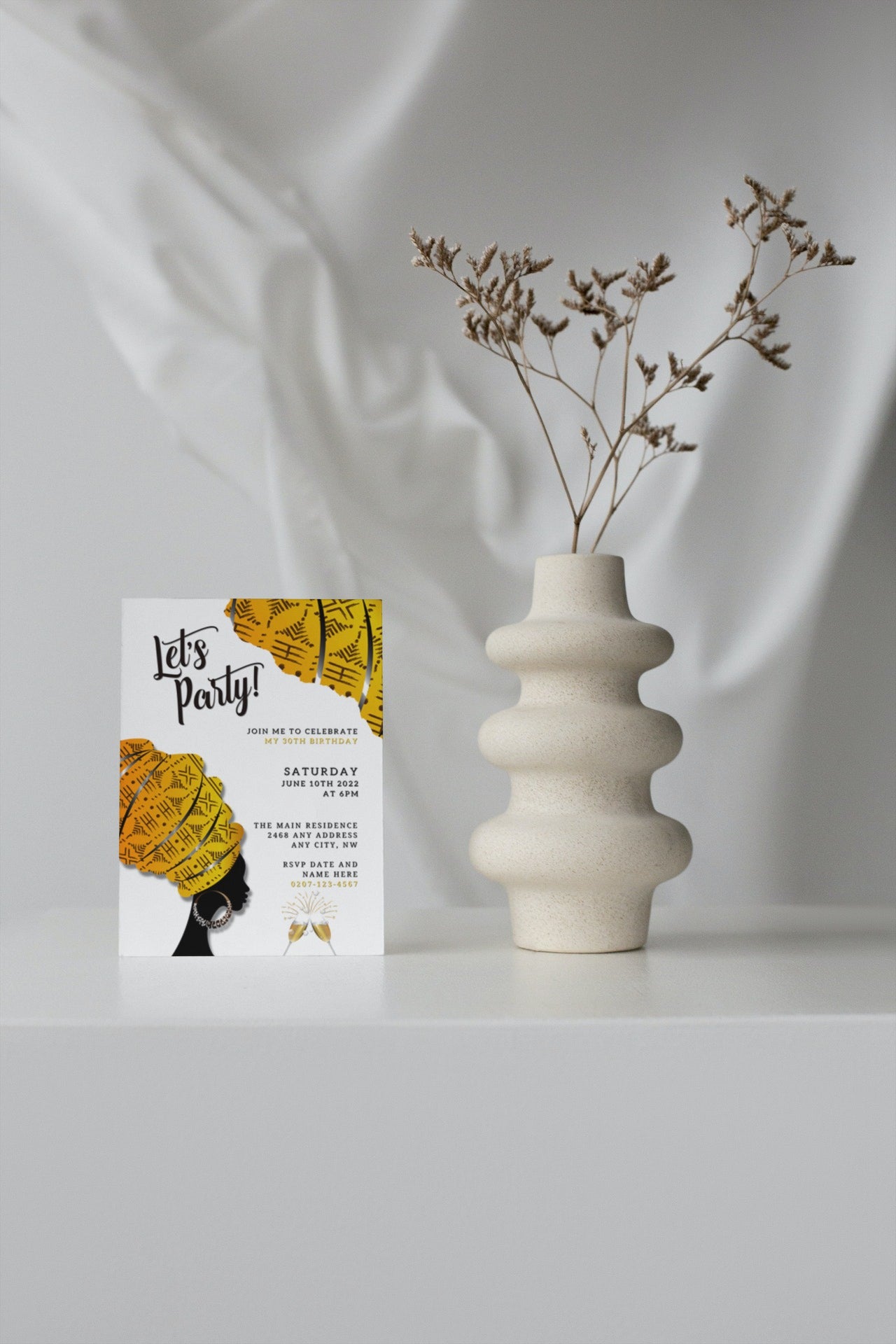 White vase with dried flowers next to a yellow and black invitation card featuring a silhouette of an African woman in a yellow turban.