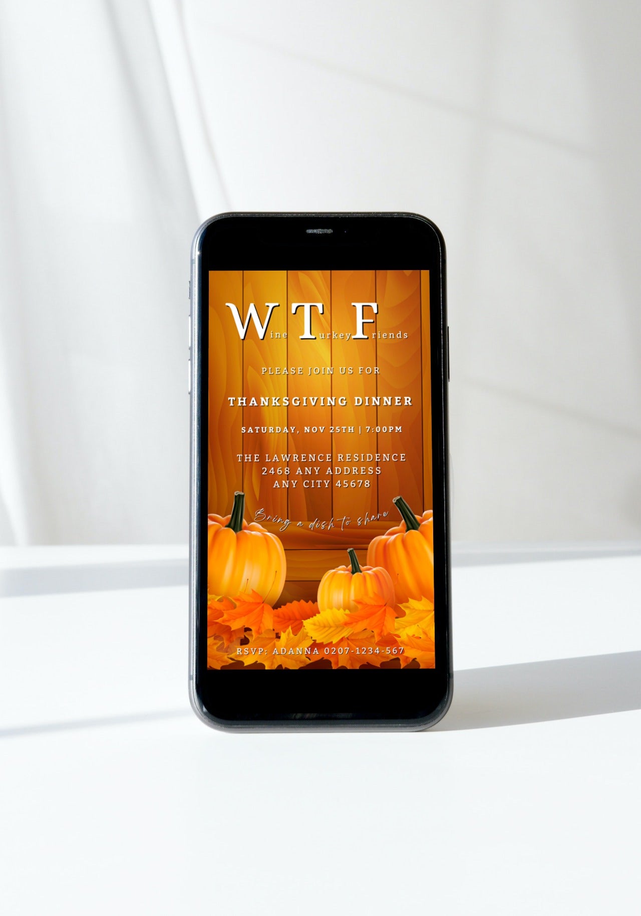 Cell phone displaying Yellow Wood Pumpkin Leaves | WTF Thanksgiving Evite template, surrounded by pumpkins and autumn leaves on a wooden background.