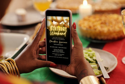 A person holding a phone displaying the Black Neon Gold Balloons | Birthday Weekend Evite template from URCordiallyInvited.