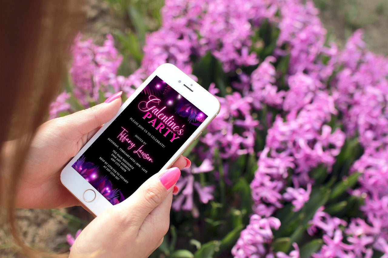 Hand holding a phone displaying a customizable Purple Pink Neon Palm Leaves Galentines Party Evite from URCordiallyInvited, with purple flowers in the background.