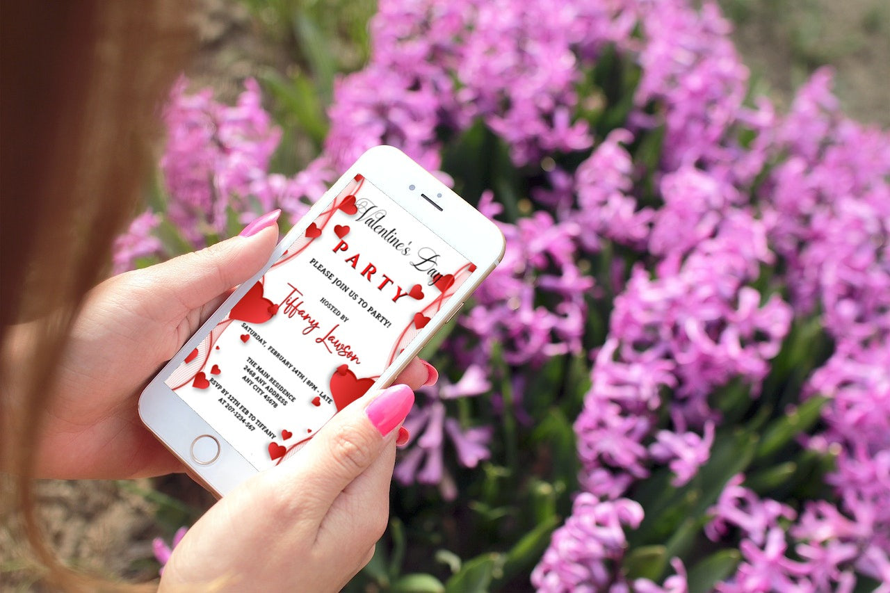 Hand holding a smartphone displaying a customizable Valentine's Party Evite with white red border hearts, designed for editing and sharing digitally via Canva.