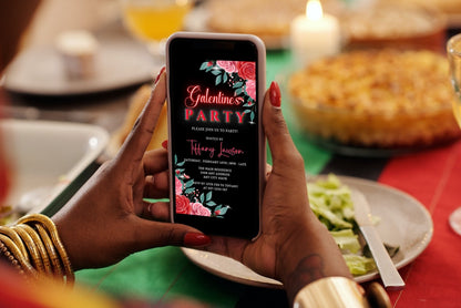 Person holding a phone displaying the Red Teal Floral Rose Galentines Party Evite, a customizable digital invitation template from URCordiallyInvited.