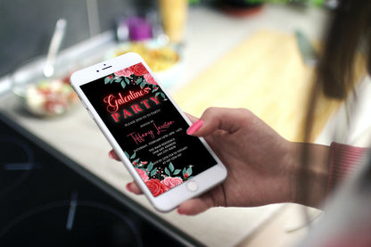 Hand holding a phone displaying a Red Teal Floral Rose Galentines Party Evite, customizable via Canva for easy sharing.