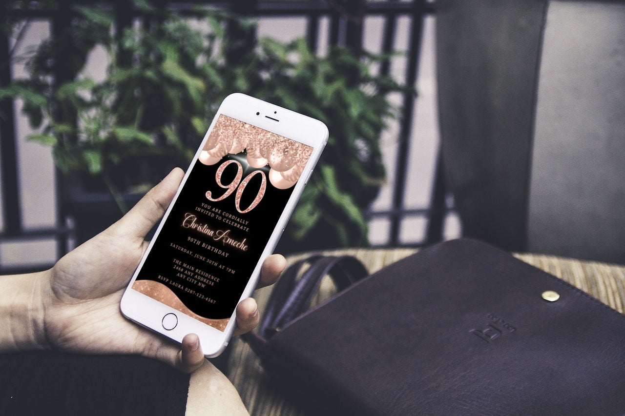 Hand holding a smartphone displaying the customizable Rose Gold Balloons Glitter 90th Birthday Evite from URCordiallyInvited, editable via Canva and shareable digitally.