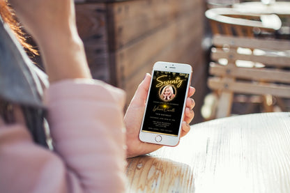 A person holding a smartphone displaying a customizable Digital Gold Neon W/Oval Photo Frame for a 70th Birthday Evite, ready for personalization using Canva.