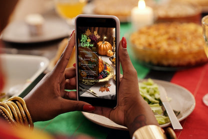 Thanksgiving dinner video invite on phone screen, featuring Let's Gather with turkey, pumpkins, and leaves, held by someone at a festive table.