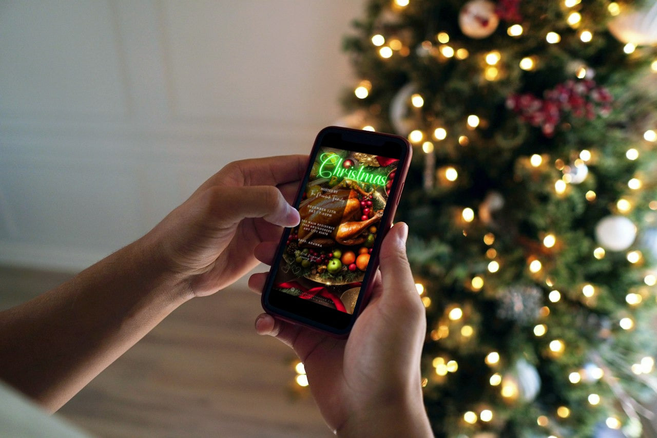 Person holding a phone displaying a Green Neon Festive Food Platter | Christmas Video Invitation with a decorated Christmas tree in the background.