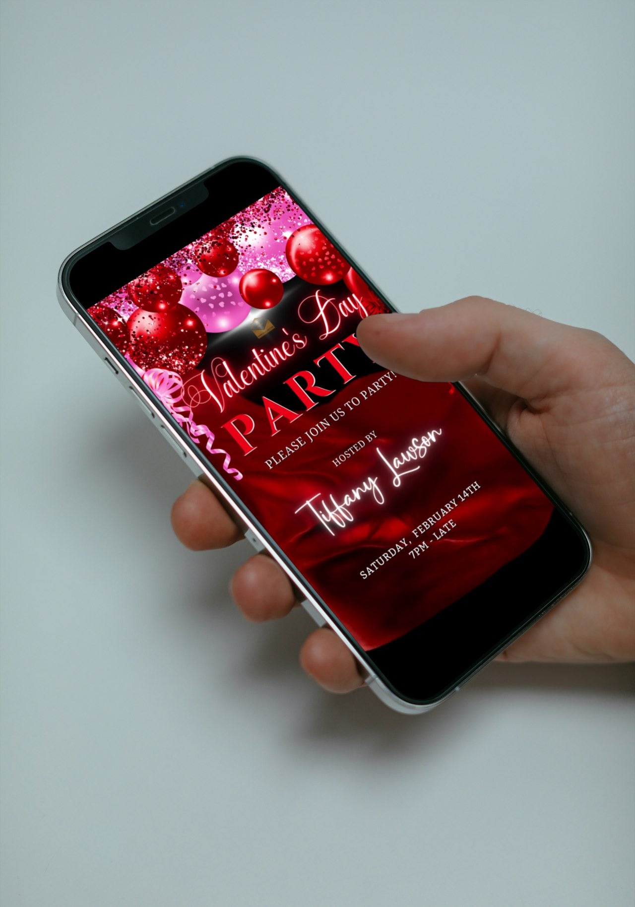 Hand holding a smartphone displaying a customizable digital invitation for Valentine's Day with pink and red silk neon balloons.