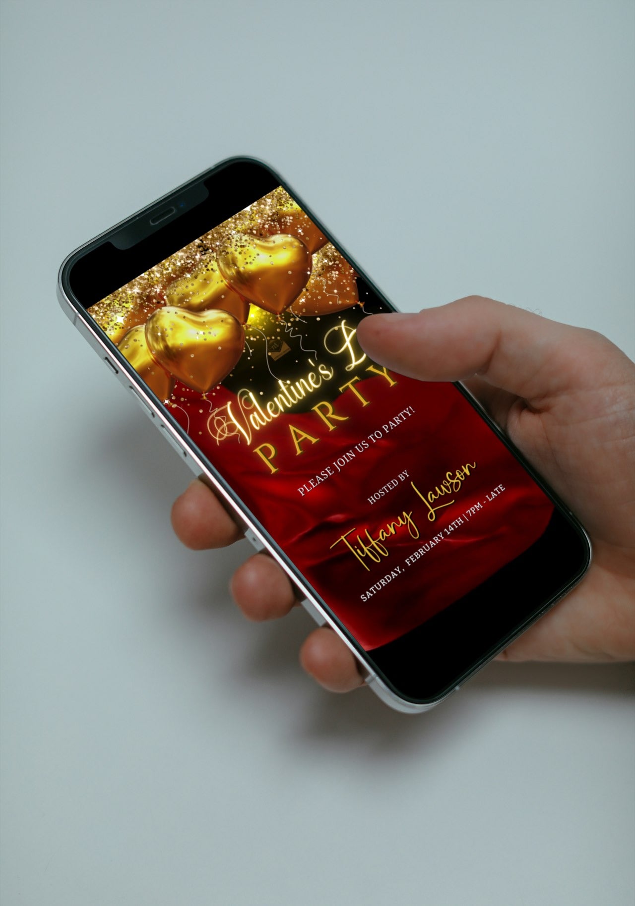 Hand holding a smartphone displaying a customizable digital invitation featuring red silk, neon gold heart balloons for Valentine's Day.